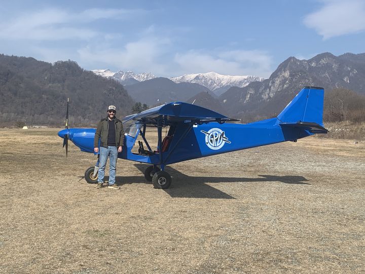 Pilota in piedi accanto al Savannah blu del GPV sul prato del campo di volo, con le montagne innevate sullo sfondo